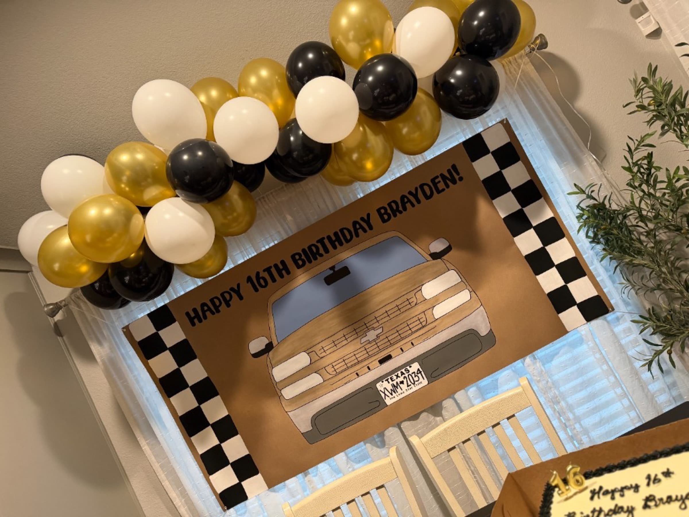 Kraft paper banner hanging at a party with gold, black, and white balloons, reading Happy 16th Birthday Brayden with a painted gold truck and checkered borders